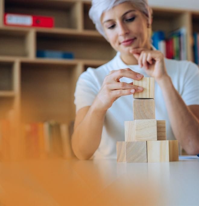 A young woman with short white hair places a wooden block on a tower made of six wooden blocks. A bookshelf can be seen in the background.