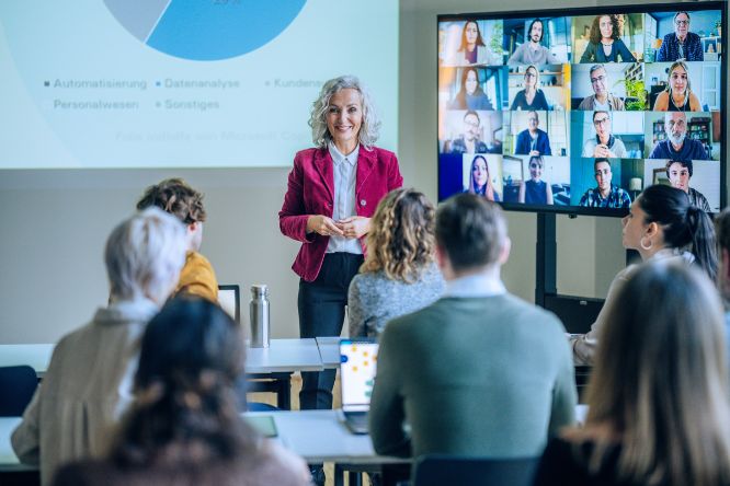 Students in a course with their backs to the camera. In front is a lecturer talking to the students. Behind her is a large screen with participants in an online meeting.
