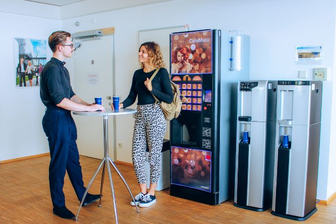 Ein Student und eine Studentin stehen an einem Stehtisch vor dem Kaffee- und Wasserautomaten