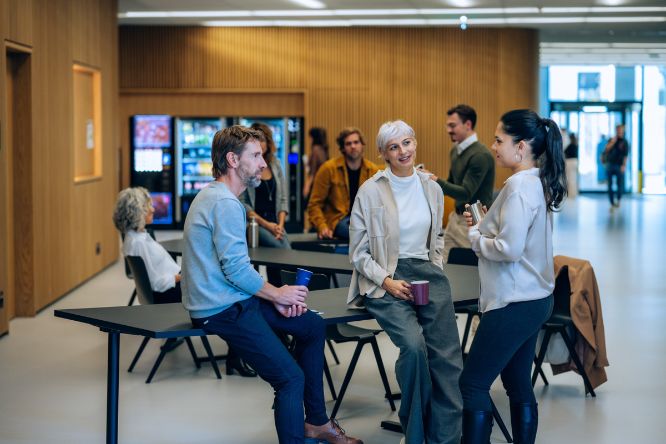 Studierende, angelehnt an Tischen, unterhalten sich im Foyer des Traktes U. Im Hintergrund sind Kaffee- und Getränkeautomaten zu sehen.
