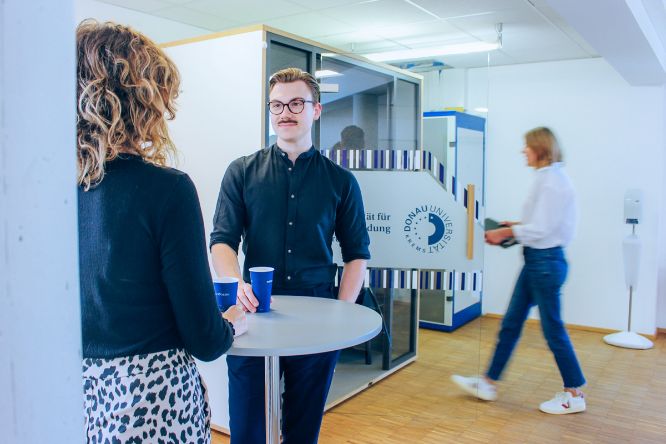 Eine Studentin und ein Student stehen am Stehtisch zusammen und unterhalten sich. Im Hintergrund geht eine Studentin in die Akustikkabine rein.