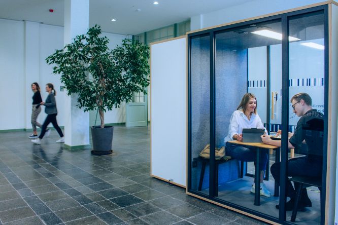 Eine Studentin und ein Student sitzen in der Akustikkabine und arbeiten. Zwei Studierende gehen im Hintergrund vorbei.