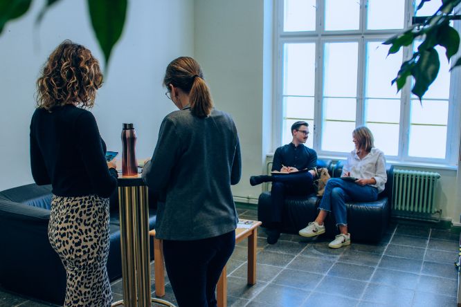 Zwei Studentinnen stehen am Stehtisch mit dem Rücken zur Kamera. Ein Student und eine Studentin sitzen im Hintergrund auf den Couch-Möbeln und unterhalten sich.