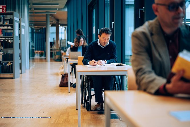 Studierende an den Leseplätzen in der Bibliothek im Trakt C Erdgeschoss, ein Student im Rollstuhl