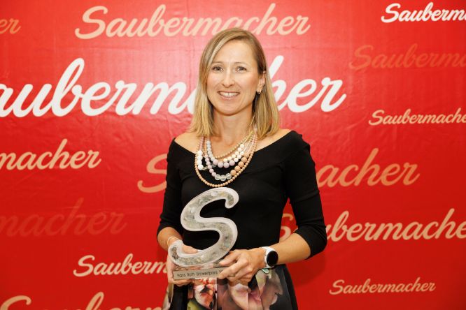 Frau mit blonden Haaren, schwarzem Oberteil und Pokal in Händen vor roter Logowand