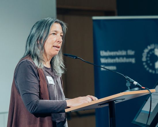 Frau mit grauen, glatten, schulterlangen Haaren und mauvem Outfit hält Vortrag.