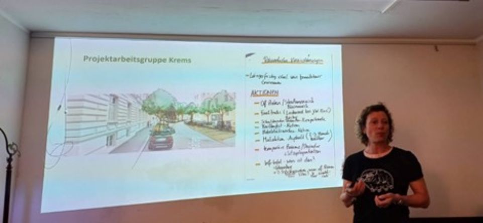 Woman talks in front of a picture of a chart showing a street with parking cars and trees
