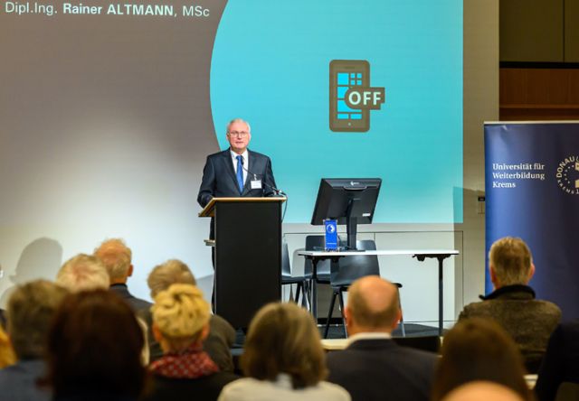 Mann in blauem Anzug vor Publikum und blauer Leinwand mit dem Zeichen "Handys Off"