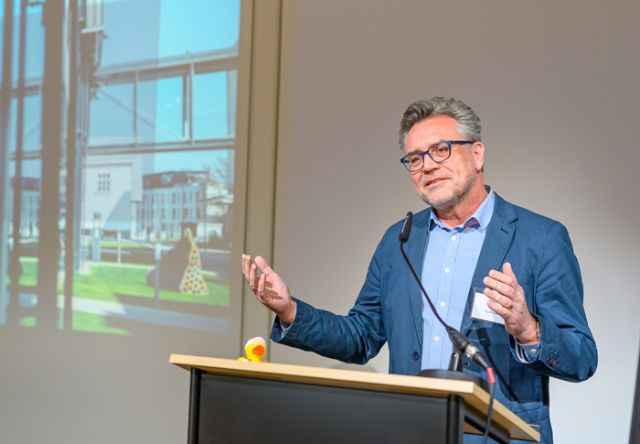 Mann mit blauem Anzug und hellblauem Hemd und Brille spricht in Schwanenhalsmikrophon