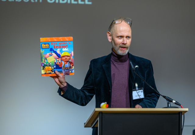 Mann in schwarzem Sakko und violettem Rollkragenpullover hält Buch von Bob, dem Baumeister in die Höhe