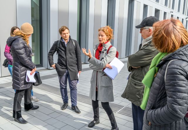 Dame in grauem Mantel und rotem Schal erklärt Umstehenden auf Betonfläche etwas