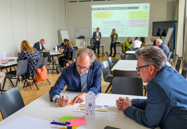 Personen sitzen in Gruppen um Tische und schreiben, diskutieren