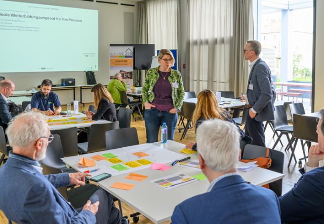 Personen sitzen in Gruppen um Tische und schreiben, diskutieren
