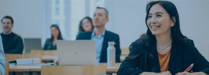 Studierende hören einer vortragenden Person in einer Lehrveranstaltung zu. Im Vordergrund sitzt eine junge, asiatische Frau, die lächelt.