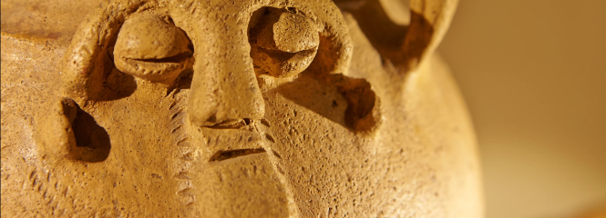 A close-up of a clay vessel with a sculpted human-like face featuring deep-set eyes, a long nose, and a textured surface.