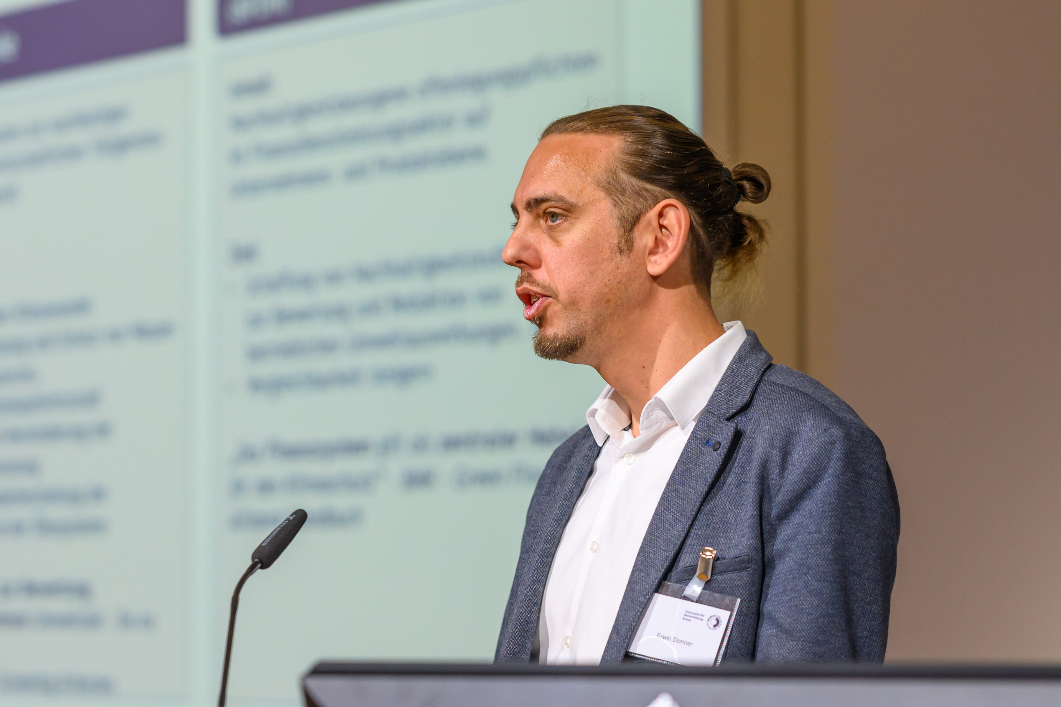 Mann mit Dutt und Bärtchen im grauen Anzug mit weißem Hemd seitlich vor weißem Hintergrund mit blauer Schrift