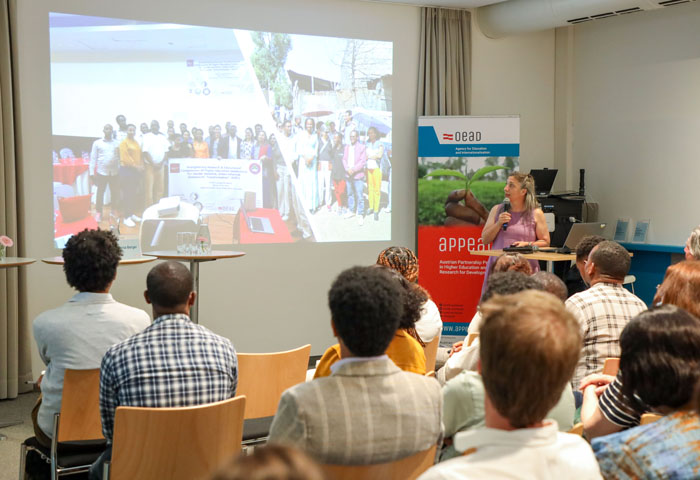 © OeAD/Nina Zuckerstätter Lady in a purple dress stands in front of an audience with a microphone and gives a lecture
