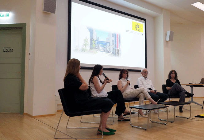 5 Personen sitzen auf Sesseln vor Beamerleinwand und diskutieren