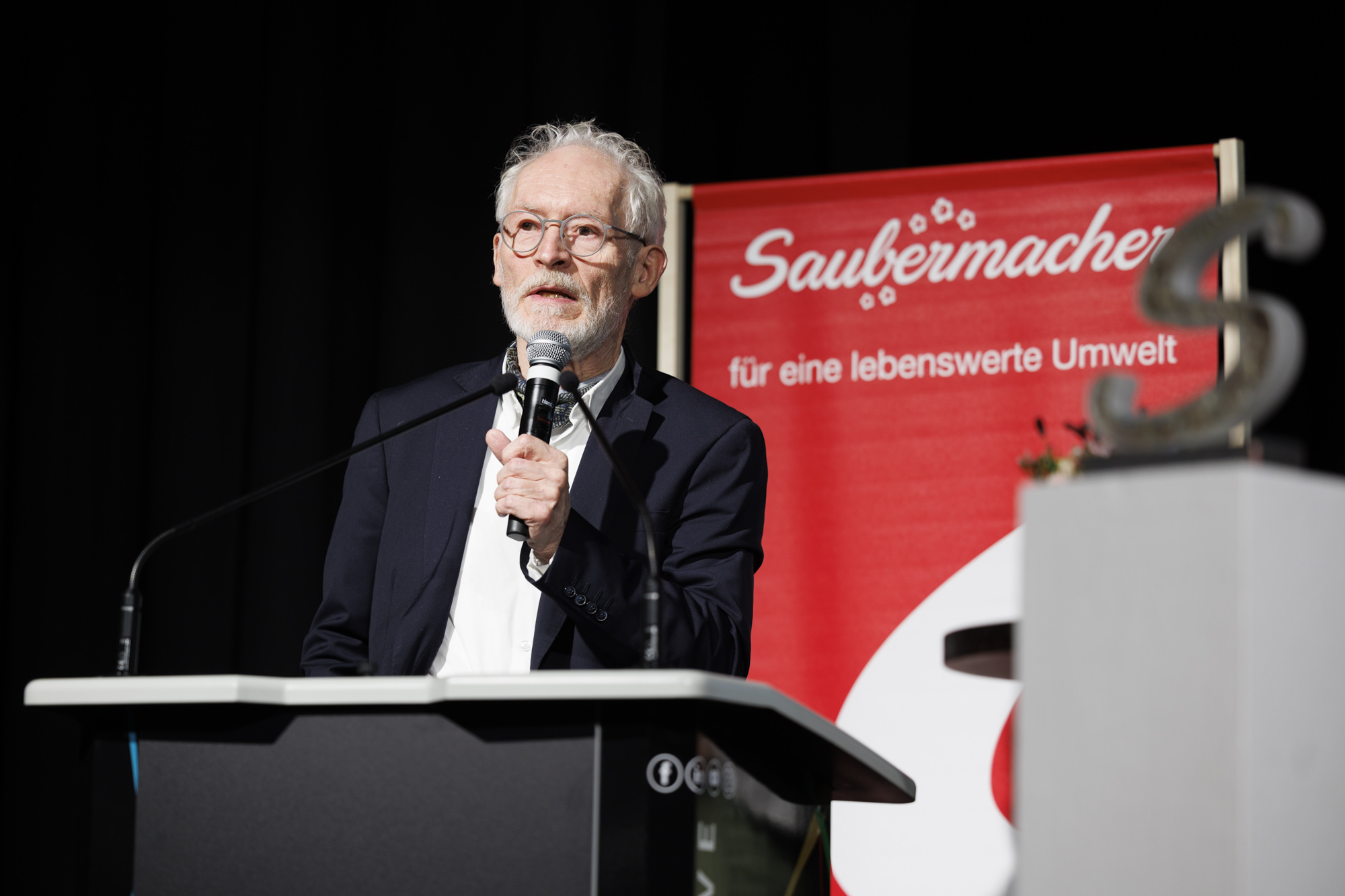 Mann mit weißen Haaren und Bart und Brille in Anzug mit Mikrofon in der Hand