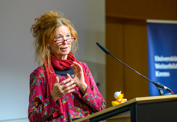 Frau mit roten hochgesteckten Haaren, rotem Shirt, rotem Schal um den Hals und Lesebrillen hält Vortrag