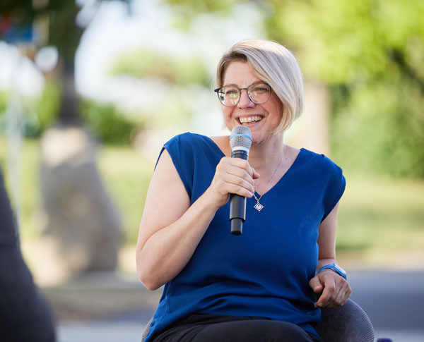 sitzende Dame mit Brille, einem Lächeln und blauem T-Shirt hat Mikro in der Hand