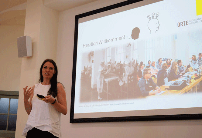 Dame mit langen schwarzen Haaren und weißer Bluse steht vor Beamerleinwand mit Präsentation