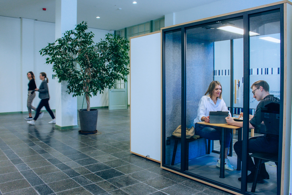 © Stefanie Gilber Eine Studentin und ein Student sitzen in der Akustikkabine und arbeiten. Zwei Studierende gehen im Hintergrund vorbei.