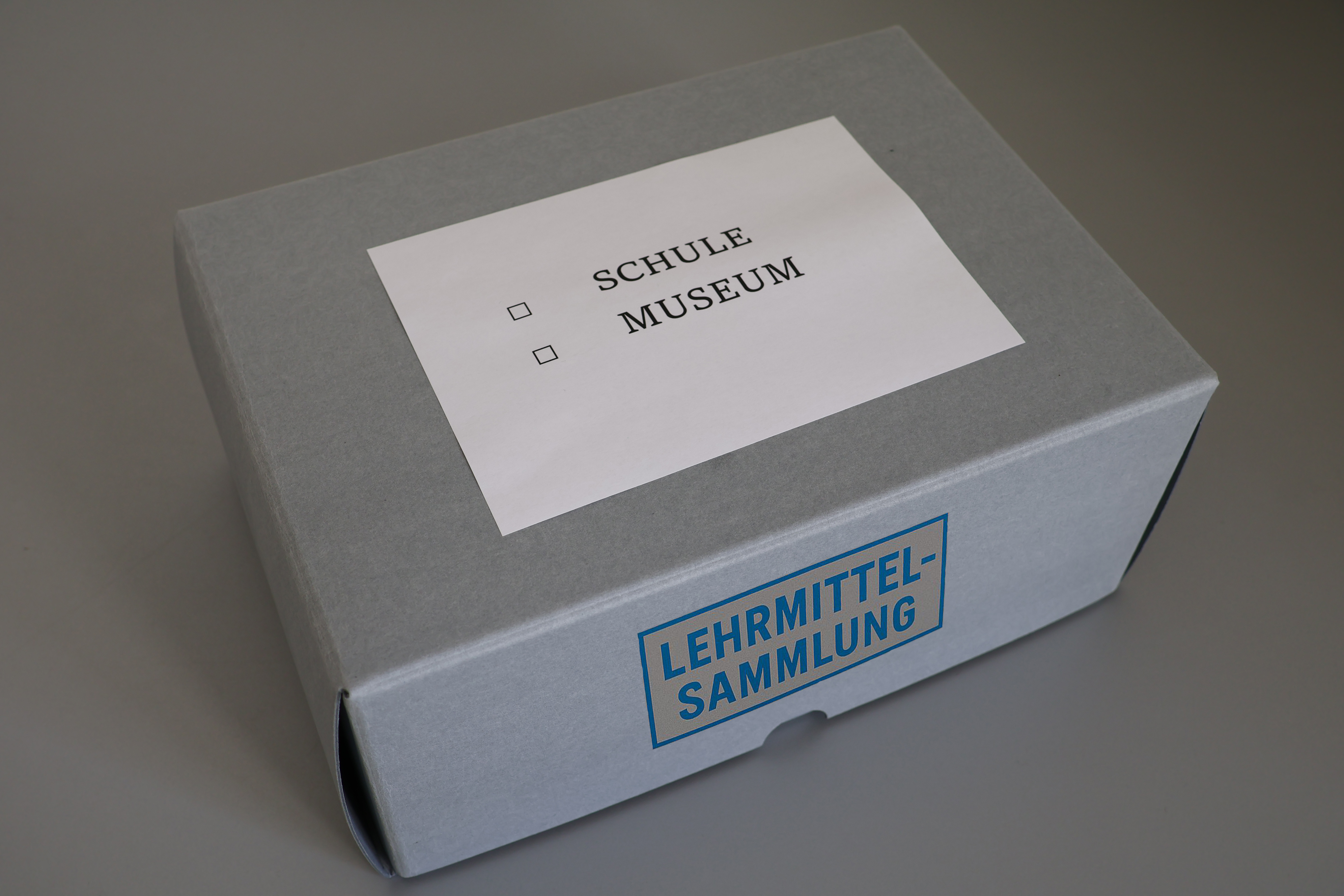 Im Zentrum steht eine verschlossene Box aus säurefreiem Karton. Auf dem Deckel klebt ein Zettel auf dem steht zum Ankreuzen Schule und Museum. Auf der Seite ist ein Aufkleber mit der Aufschrift Lehrmittelsammlung.