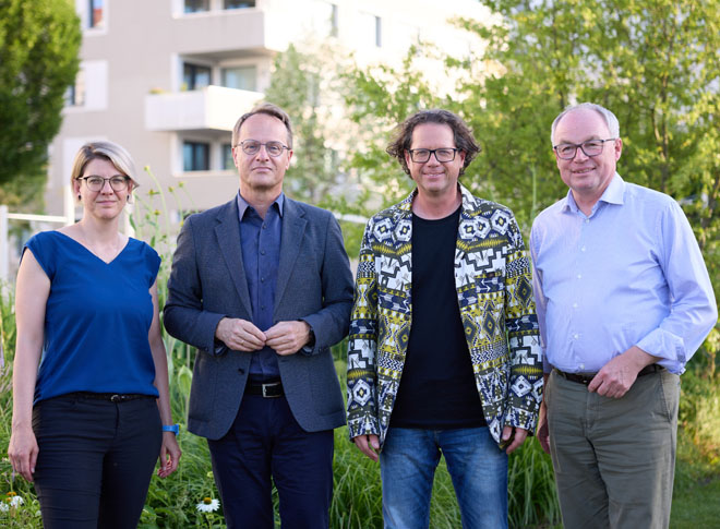 GRuppenfoto mit Dame in blauem T-Shirt und Brille, Herr in blauem Sakko und Brille, Herr mit buntem Sakko, Brille und dunklen Locken und Herr mit hellblauem Hemd und Brille