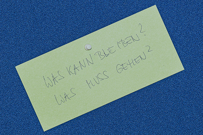 Gelbe Moderationskarte auf blauer Pinnwand mit der Aufschrift "Was kann bleiben? Was muss gehen?"