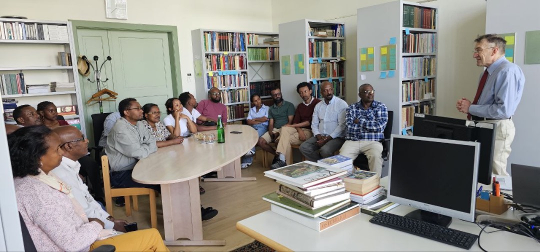 © DBU/Elisabeth Huber People in a semicircle listen to a man with glasses, a red tie, and a blue shirt in a room with many bookshelves during a lecture.