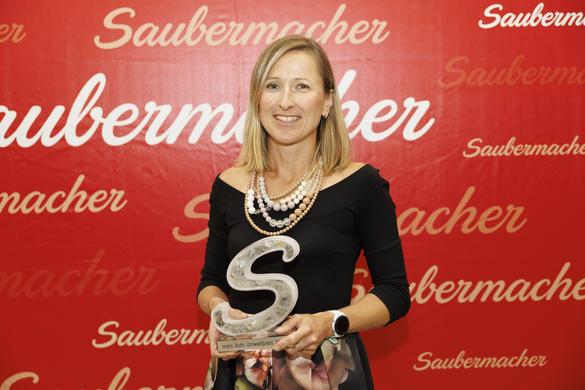 Frau mit blonden Haaren, schwarzem Oberteil und Pokal in Händen vor roter Logowand