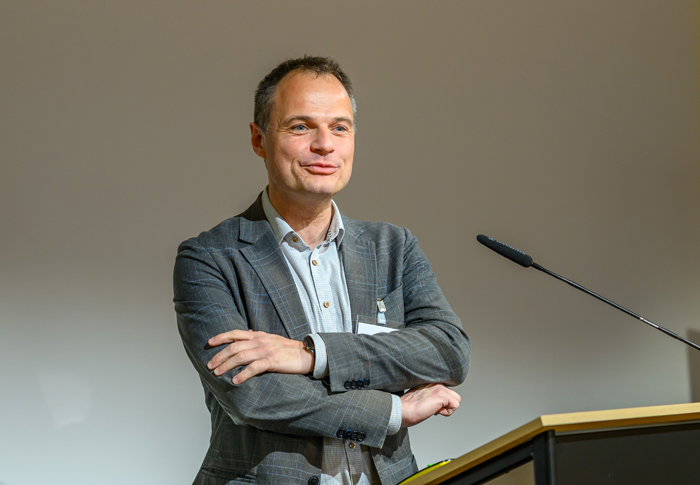 Mann in grauem Anzug und hellem Hemd mit verschränkten Armen bei Rednerpult und vor Schwanenhalsmikrophon