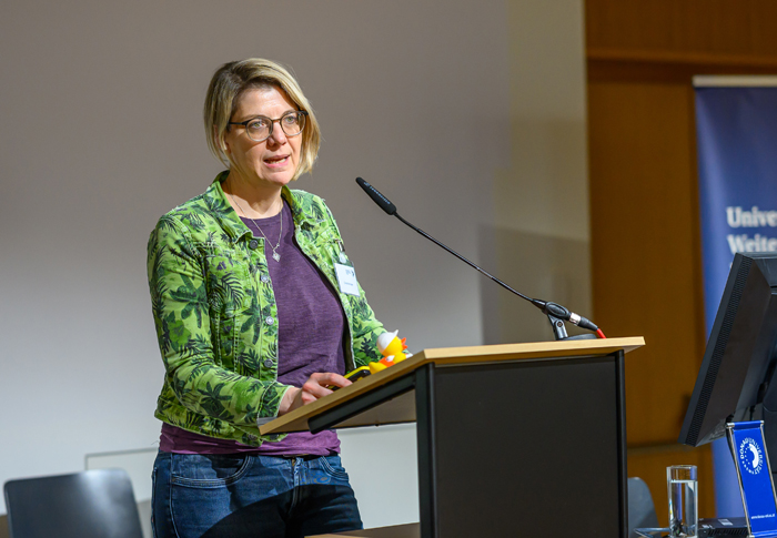 Frau mit grünem Blazer mit Blättern und violettem Shirt und Brille hält einen Vortrag