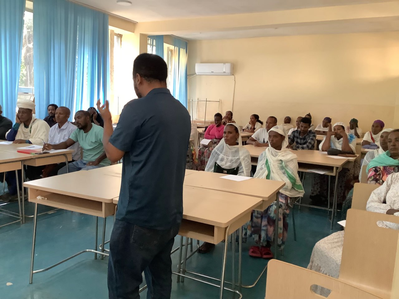 Mann mit blauem Outfit spricht vor afrikanischen Frauen, die in Raum auf Schulbänken sitzen