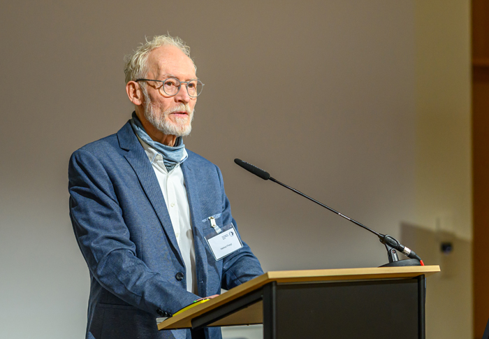 Mann mit weißem lockigen Haar, Bart und Brille bei Rednerpult mit Schwanenhalsmikrophon