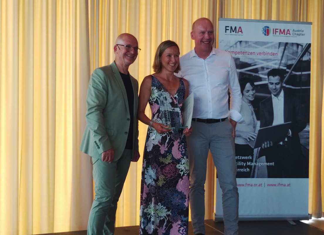 Dame in Blumenkleid flankiert von 2 Herren bei Preisverleihung